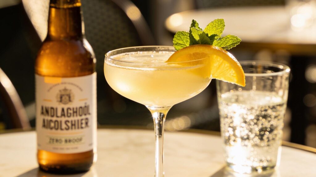 A zero-proof cocktail in a coupe glass with citrus and mint on a sunlit marble cafe table, a non-alcoholic beer softly blurred behind.